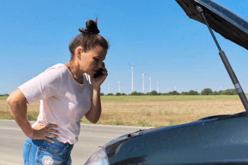 How We Can Prepare Your Car for the Summer Heat with Repair One in The Woodlands Tx and Klein Tx image of middle aged woman with hair in bun in summer clothes on the cell phone with hood open on car on the side of the road