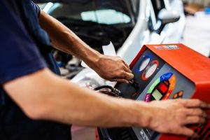 Can I Perform A/C Recharge Service at Home? | Repair One at the Woodlands. Closeup image of a mechanic’s hands refilling a car’s air conditioner with a refrigerant refilling machine.
