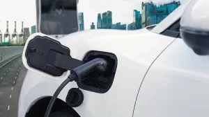 Do Hybrid & Electric Vehicles Require Special Maintenance? | Repair One in The Woodlands, TX. Image of a white electric vehicle recharging at charging station on the freeway surrounded by tall skyscrapers.