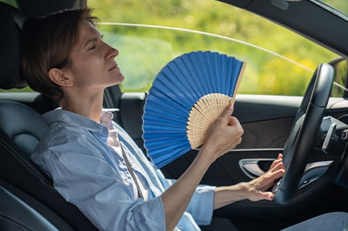 Overheated exhausted middle aged woman driver with hand fan suffering from heat in car. Concept image of “Keep Your Car Cool With AC & Cooling System Services” | Repair One in Spring/Klein, TX.