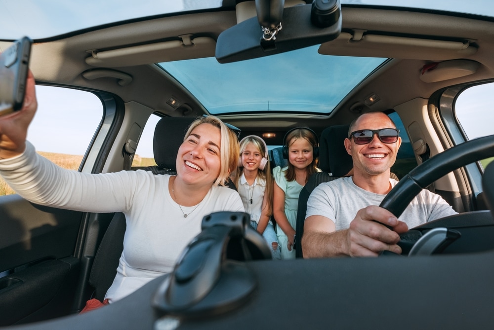 Holiday Vehicle Maintenance | Repair One The Woodlands, TX | Auto Care Services. Happy young couple with two daughters driving on road trip taking family selfie